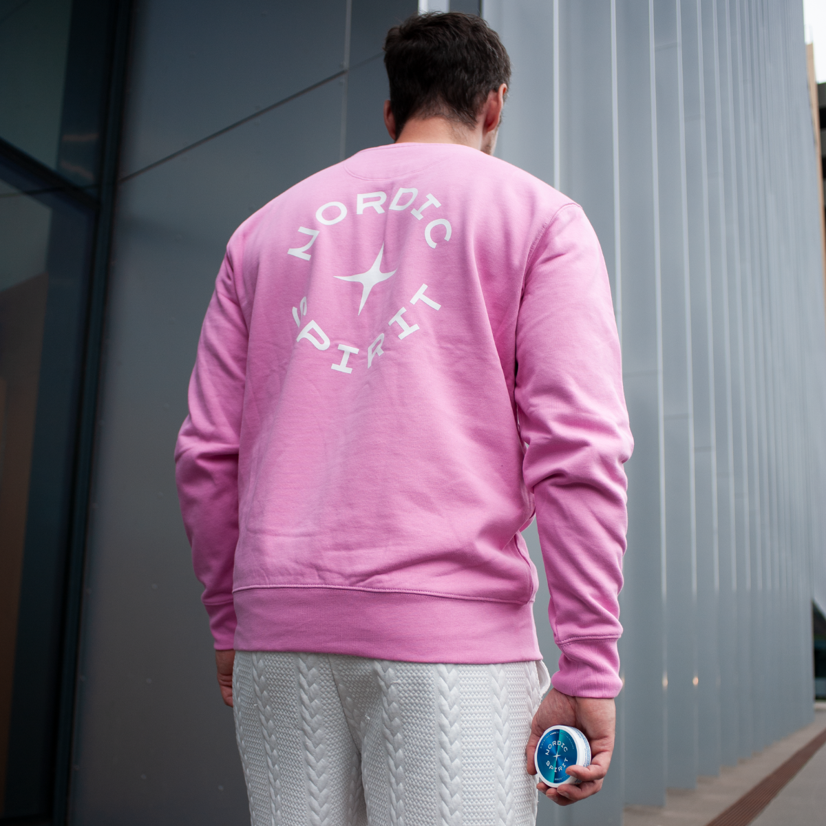 Person in einem pinken Sweatshirt mit Nordic Spirit Logo, hält eine kleine runde Dose in der Hand, vor einem modernen Hintergrund mit Glas- und Metallelementen.
