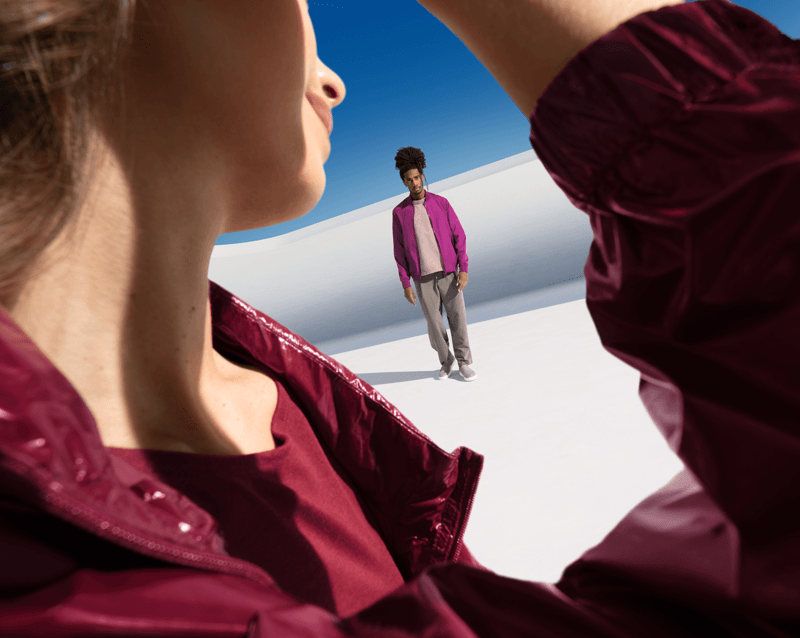 Zwei Personen in winterlicher oder sandiger Umgebung unter klarem Himmel, eine in pinker Jacke im Hintergrund, eine in bordeauxfarbener Kleidung im Vordergrund.