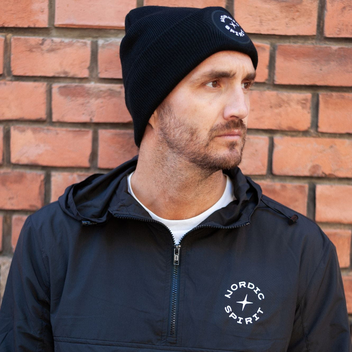 Person mit schwarzer Nordic Spirit Mütze und schwarzer Jacke vor einer Backsteinwand.