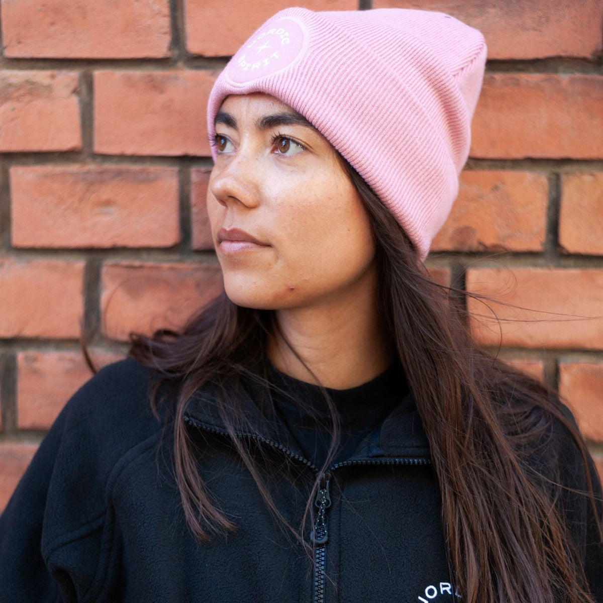Person mit rosa Nordic Spirit Mütze und schwarzer Fleecejacke vor einer Backsteinwand.
