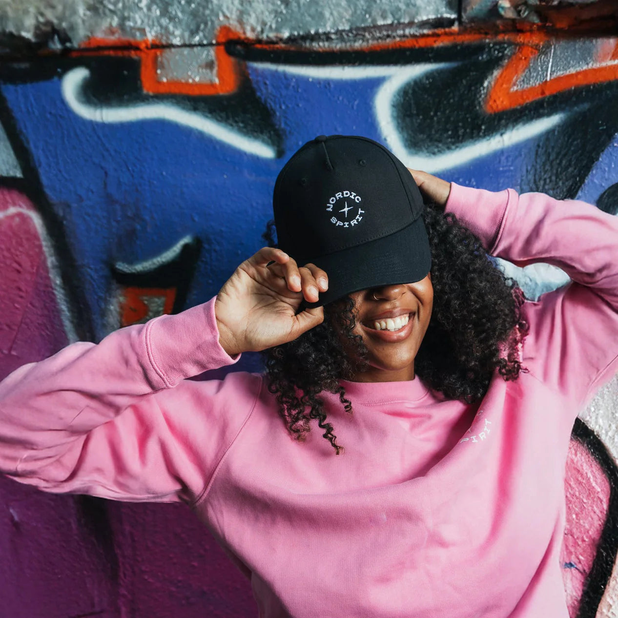 Junge Frau mit aufgesetzter Nordic Spirit Kappe posiert vor Wand mit Grafitti