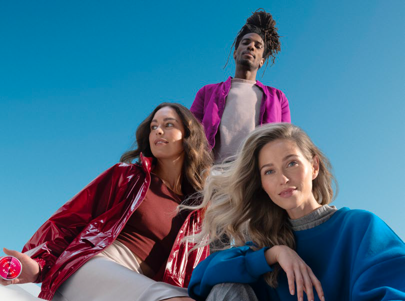 Drei Personen im Freien vor klarem blauen Himmel; die mittlere Person trägt eine pinke Jacke, links eine rote Jacke, rechts ein blaues Oberteil.