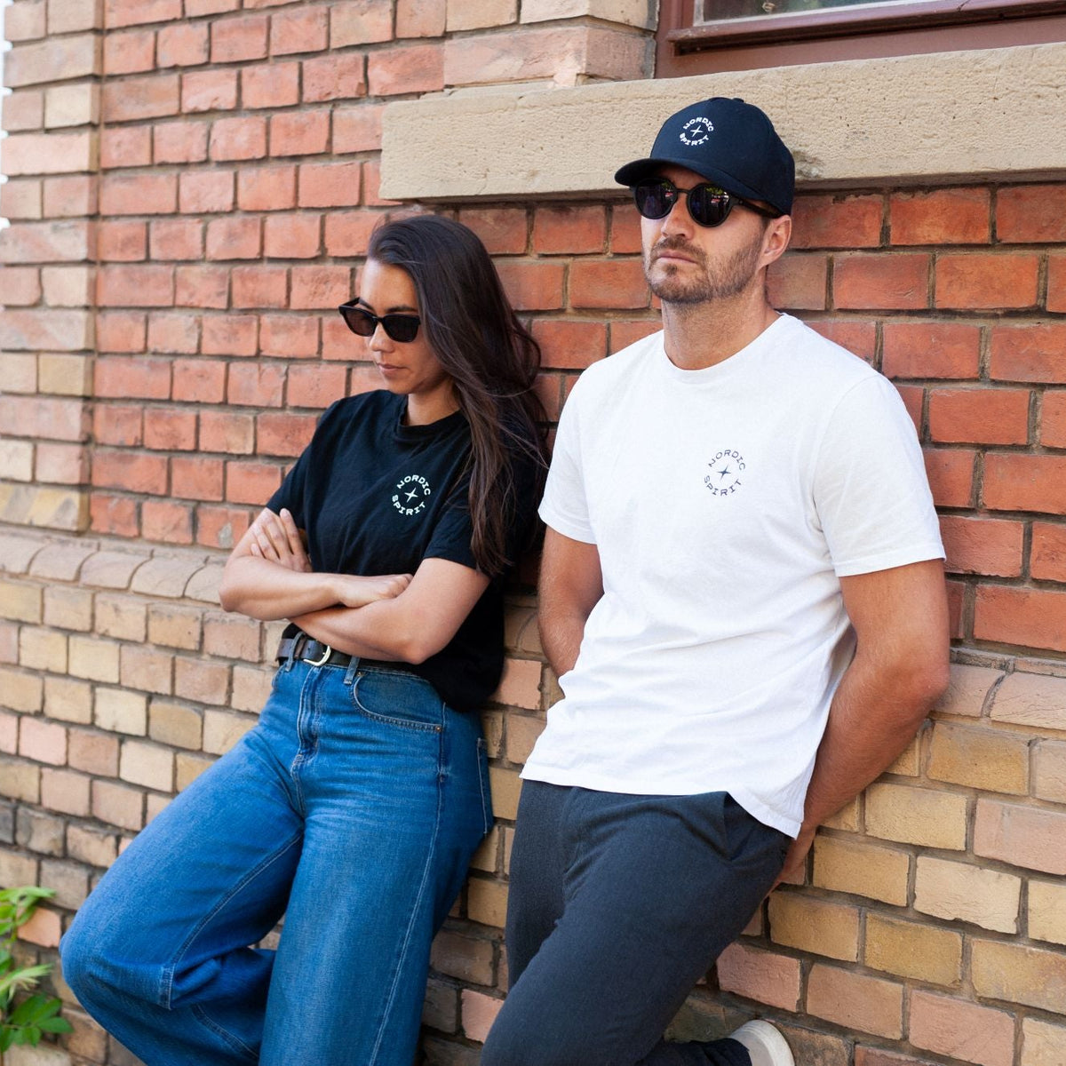 Zwei Personen mit Sonnenbrillen in schwarzen und weißen Nordic Spirit T-Shirts vor einer Backsteinwand.